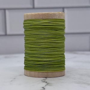 May include: A spool of olive green thread. The thread is tightly wound around a cardboard core. The background is a white and gray tiled wall and a white marble surface.