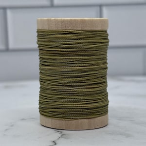 May include: A cylindrical wooden spool wrapped with olive green thread. The spool features a natural light-coloured wood grain on the top and bottom circular ends, while the centre is densely wound with thin, uniform olive green cord.