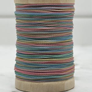 May include: A wooden spool wrapped with multi-colored thread in pastel shades of pink, blue, green, and yellow. The thread is wound tightly around the cylindrical wooden core, creating a layered, striped appearance with a smooth, uniform texture.