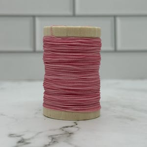 May include: A spool of pink thread. The thread is tightly wound around a light-coloured wooden core. The thread is a solid, medium pink colour. The spool is on a white and grey marble surface.