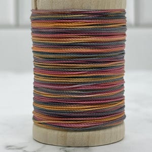 May include: A wooden spool wrapped with variegated thread featuring a color palette of muted orange, dusty rose, and charcoal gray. The thread is wound tightly around the cylindrical wooden core, creating a layered, multi-colored horizontal striped effect.