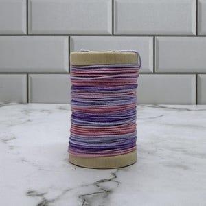 May include: A wooden spool wrapped with thin cord in alternating horizontal bands of light pink, lavender, and deep purple. The spool has a cylindrical shape with flat circular ends and is wound tightly with the multi-colored string.