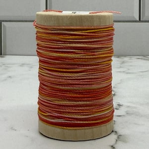 May include: A spool of colorful thread in shades of red, orange, and yellow. The thread is tightly wound around a light brown cardboard core. The colors create a gradient effect, suitable for various crafting projects.