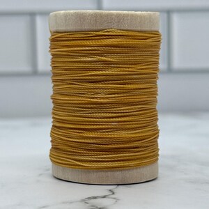 Könnte beinhalten: Eine Spule goldgelben Fadens. Der Faden ist eng um einen hellfarbigen Holzkern gewickelt. Der Faden scheint aus gewachstem Leinen oder einem ähnlichen Material zu bestehen, das für verschiedene Bastelprojekte geeignet ist. Der Hintergrund ist eine weiße Fliese.