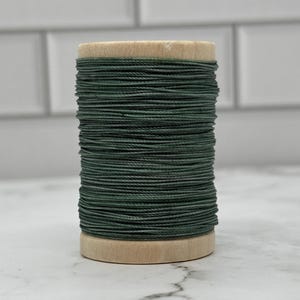 May include: A spool of dark green thread. The thread is tightly wound around a light-coloured wooden spool. The thread appears to be a solid colour, and the spool is sitting on a white surface.