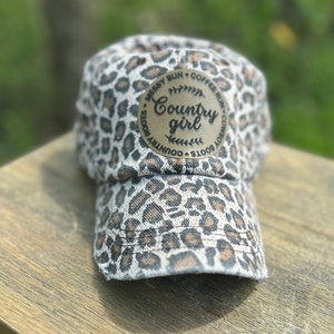 May include: A brown and white leopard print baseball cap with a brown leather patch that says "Country girl" in a white font. The patch is surrounded by a circle of text that says "Messy Bun, Coffee Runs, Cowboy Boots, Country Roots".
