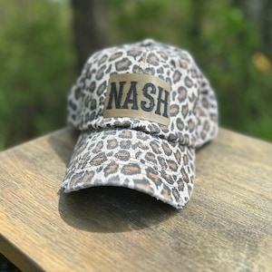 May include: A brown and white leopard print baseball cap with a brown patch that says "NASH" in black letters.