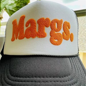 May include: A trucker hat featuring a white front panel with the word Margs. embroidered in raised orange textured lettering. The hat has a black brim and a black mesh back, with a thin black cord detail separating the front panel from the brim.