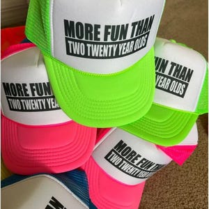 May include: A collection of trucker hats in various bright colors, including neon green, pink, and blue. Each hat has a white front panel with the text "MORE FUN THAN TWO TWENTY YEAR OLDS" printed in black.
