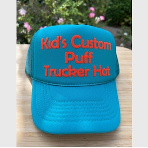 May include: A teal and orange trucker hat with the text "Kid's Custom Puff Trucker Hat" in orange puff lettering. The hat has a mesh back and a curved brim. The hat is sitting on a wooden surface.