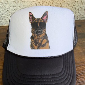 May include: Black and white trucker hat featuring a graphic of a German Shepherd wearing sunglasses. The dog is brown, black, and tan. The hat has a black brim and mesh back, with a white front panel.