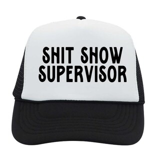 May include: A black and white trucker hat with the words "SHIT SHOW SUPERVISOR" printed in bold, black letters across the front. The hat has a white front panel and a black mesh back.