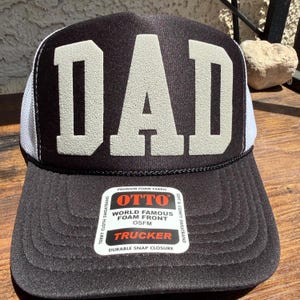 May include: A black trucker hat with white mesh side panels and a large, textured white DAD graphic on the front foam panel. A rectangular sticker on the brim features the text OTTO, WORLD FAMOUS FOAM FRONT, OSFM, TRUCKER, and TERRY CLOTH SWEATBAND.