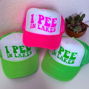 May include: Three trucker hats with the phrase "I PEE IN LAKES" in neon colors. One hat is bright pink, and two are neon green. The hats have white fronts and mesh backs. A small potted plant is in the background.