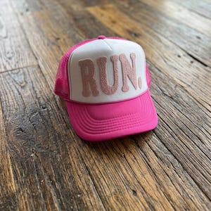 May include: A bright pink trucker hat featuring a white front panel with the word RUN. in textured, glittery pink letters. The hat has a mesh back, a solid pink brim, and a structured foam front panel, providing a classic athletic headwear look.