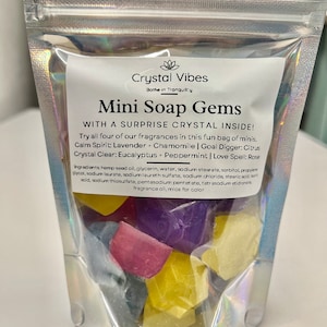 May include: A clear plastic bag filled with colorful mini soap gems shaped like crystals. The bag has a label that reads "Crystal Vibes" and "Mini Soap Gems with a surprise crystal inside!" The label lists four different scents: Calm Spirit Lavender + Chamomile, Goal Digger: Citrus, Crystal Clear: Eucalyptus + Peppermint, and Love Spell: Rose.