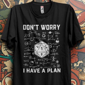 Può includere: T-shirt nero con la scritta "DON'T WORRY I HAVE A PLAN", equazioni matematiche e un dado D20. La maglietta è appesa a una gruccia nera su uno sfondo colorato.