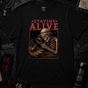 Puede incluir: Una camiseta negra con un estampado gráfico de un esqueleto con casco colonial que observa con una lupa a un escarabajo sobre un tronco. El texto "STAYING ALIVE" aparece impreso en rojo sobre la ilustración dentro de un marco rectangular.