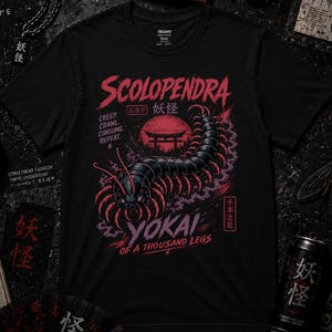 Può includere: T-shirt nera con grafica di millepiedi rossa e nera e la scritta "Scolopendra Yokai of a Thousand Legs". Il design include testo giapponese e un torii rosso. Sono visibili vari oggetti a tema giapponese.