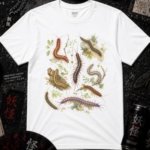 May include: A white short-sleeved t-shirt featuring a detailed, colorful botanical-style illustration of various centipedes and millipedes arranged in a cluster, surrounded by small green leaves and sprigs of foliage on the front center.