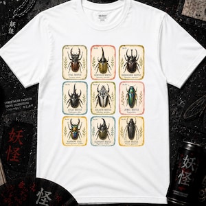 Könnte beinhalten: Ein weißes Kurzarm-T-Shirt mit einem grafischen Aufdruck von neun verschiedenen Käferarten, die in einem Raster angeordnet sind. Jeder Käfer ist von einem farbigen rechteckigen Rahmen umgeben, mit seinem Namen und wissenschaftlichen Namen darunter.