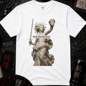 Könnte beinhalten: Ein weißes T-Shirt mit kurzen Ärmeln und einem zentralen Aufdruck einer klassischen Statue, die ein Schwert und einen abgetrennten Kopf hält. Der Text "Girls Will Be Girls" steht in einem Rechteck auf dem Torso der Statue.