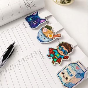 May include: Four holographic magnetic bookmarks featuring a cat head, a bowl of ramen, a superhero, and a milk carton with a face. The bookmarks are placed on a white planner page with a calendar for May 2026, a to-do list, and a time table.