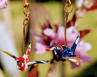 May include: A pair of gold origami crane earrings. The cranes are made of paper and are decorated with red, white, blue, and gold patterns. The earrings are hanging from gold hooks.