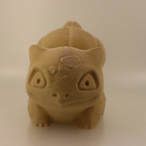 May include: A brown 3D printed figurine of a Pokemon character, resembling a small dinosaur with large eyes and a heart-shaped marking on its forehead.