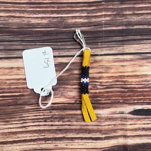 May include: A small beaded ornament with a yellow leather tassel. The beads are black, white, and blue. The ornament is attached to a white string and a tag with the number 159.