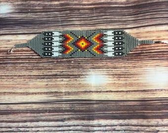 Könnte beinhalten: Ein Perlenarmband mit grauem Grund und einem geometrischen Muster in Rot, Gelb, Weiß und Schwarz. Das Armband hat eine rechteckige Form mit einem zentralen Rautenmuster, das auf einer Holzoberfläche präsentiert wird.