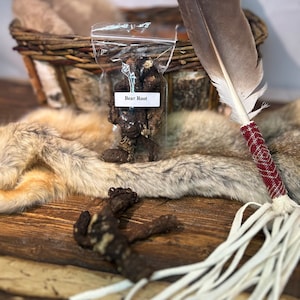 Könnte beinhalten: Ein transparenter Plastikbeutel gefüllt mit getrockneter Bärwurzel, beschriftet mit "Bear Root". Der Beutel liegt auf einer braunen Holzoberfläche mit einem weißen Fell und einer braunen Feder mit roten und weißen Perlen.