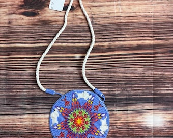 May include: A round, beaded pendant necklace with a blue background and a central starburst design in red, yellow, green, and orange. The pendant has white animal shapes and is attached to a white braided cord.