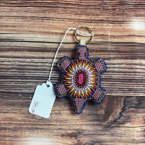 May include: A beaded turtle-shaped keychain with a silver keyring. The turtle design features a central oval of pink beads, surrounded by concentric circles of yellow, orange, and red beads. The outer edges are outlined in blue and purple beads.