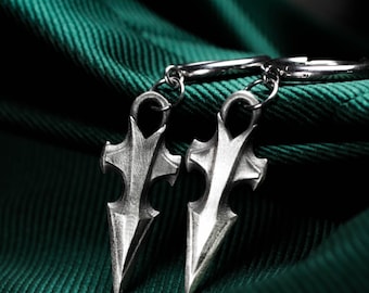 Könnte beinhalten: Ein Paar silberfarbene Creolen mit baumelnden dolchförmigen Anhängern. Jeder Anhänger hat eine spitze Form, einen Mittelsteg und symmetrische Aussparungen im oberen Bereich und ist mit einem kleinen Metallring an der Creole befestigt.