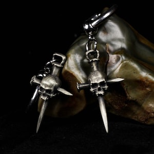 Pode incluir: Um par de brincos prateados com uma caveira com uma adaga e uma cruz. Os brincos estão pendurados em um anel prateado. O fundo é uma superfície escura e texturizada.