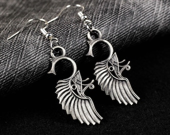 Puede incluir: Un par de pendientes colgantes de metal plateado con un diseño de ala estilizada, que presenta detalles lineales intrincados y una sección superior curva. Los pendientes cuelgan de ganchos simples de metal plateado para orejas perforadas.