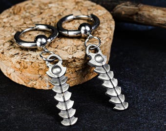 Könnte beinhalten: Ein Paar silberfarbene Creolen mit einem hängenden Fischgräten-Anhänger. Jeder Ohrring besteht aus einem runden Ring mit einer kleinen Kugel, der mit einem segmentierten, wirbelsäulenartigen Anhänger mit Metallfinish verbunden ist.