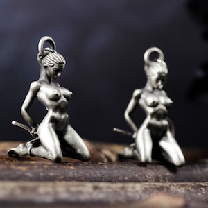 以下が含まれることがあります： 銀色のペンダント2つ。ひざまずいた女性の姿で、両手は後ろで縛られている。ペンダントの上部には吊り下げ用のループが付いている。背景は暗くぼやけたグラデーション。