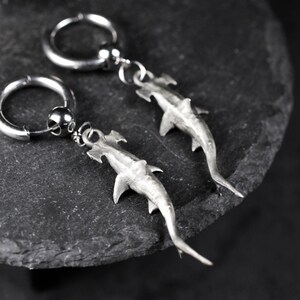 May include: A pair of silver-toned hoop earrings with dangling hammerhead shark charms. The earrings are displayed on a dark, textured surface, showcasing the detailed design of the shark pendants and the simple elegance of the hoops.