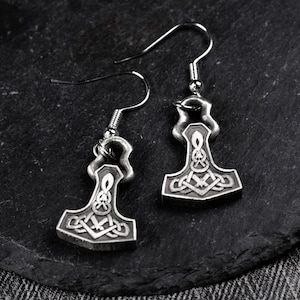 Puede incluir: Un par de pendientes plateados con diseño de martillo, con intrincados patrones de nudos celtas. Los pendientes tienen cierre de gancho y se muestran sobre una superficie oscura y texturizada. Los colgantes de martillo son detallados y simétricos.