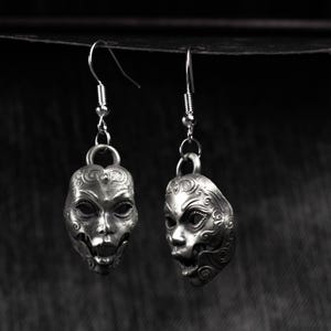 May include: A pair of silver-toned drop earrings. Each earring features a detailed mask design with intricate facial features and decorative patterns. The earrings hang from a simple hook and are suspended against a dark background.