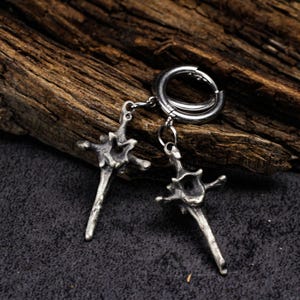 May include: Silver-coloured earrings with an abstract design. Each earring has a cross-like shape with detailed features, hanging from a circular hoop. The earrings are displayed on a dark, textured surface with a wooden background.