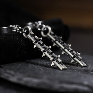 May include: A pair of silver-toned earrings. Each earring features a linear design with multiple, evenly spaced, pointed spikes. The earrings are suspended from a small, circular clasp. The background is dark, emphasizing the metallic sheen of the jewelry.