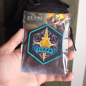 May include: A hexagonal embroidered patch featuring a yellow starburst design over a black geometric grid, bordered in teal. A teal banner across the center displays the Cyrillic text ICKPA. The patch is enclosed in a clear plastic sleeve.