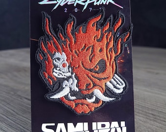 May include: An embroidered patch featuring a stylized red and orange flaming oni mask design with white fangs. The patch is mounted on a black display card with the text CYBERPUNK 2077 at the top and the word SAMURAI printed in white at the bottom.
