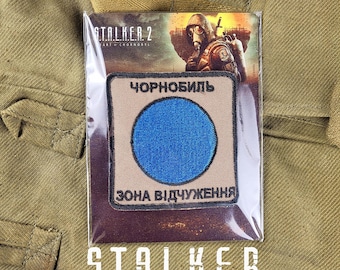 Puede incluir: Un parche marrón con el texto "STALKER 2", "ЧЕРНОБЫЛЬ" y "ЗОНА ВІДЧУЖЕННЯ" en escritura cirílica. El parche presenta un círculo azul sobre un fondo blanco.