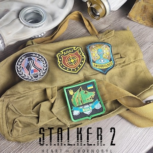 Peut inclure: Un sac en toile kaki avec plusieurs écussons brodés. Les écussons présentent divers motifs, dont un bouclier avec une cible, un atome stylisé et un loup vert. Le texte "STALKER 2 HEART OF CHORNOBYL" est affiché en bas.