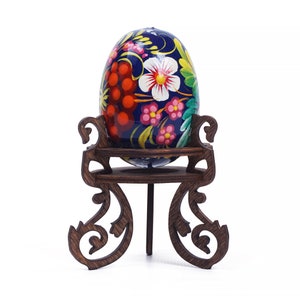 May include: A hand-painted Easter egg with floral designs in shades of red, pink, yellow, and green on a dark blue background. The egg is displayed on a wooden stand with intricate carvings.