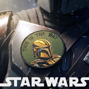 Può includere: Una toppa rotonda con la scritta "THIS IS THE WAY" che circonda un disegno di casco Mandaloriano dorato e verde. La toppa è fissata su una superficie scura e strutturata, con il logo "STAR WARS" in basso.